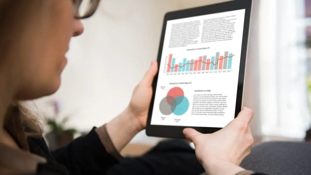 Woman is sitting in a living room looking at a tablet computer showing data, statistics and graphs.